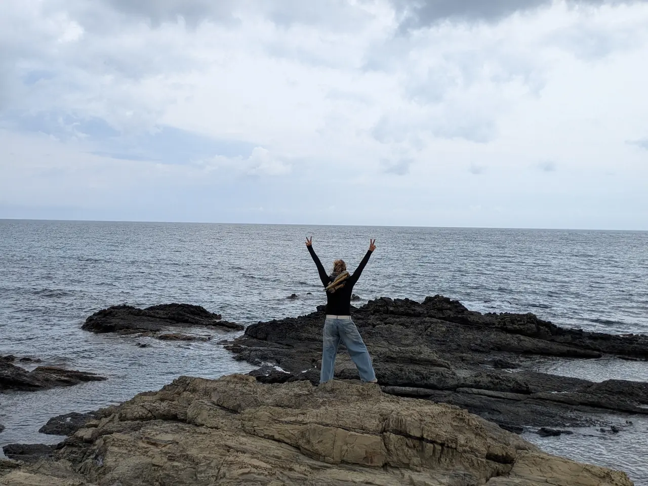 Una persona amb els braços alçats davant del mar, sobre unes roques, en un dia ennuvolat.